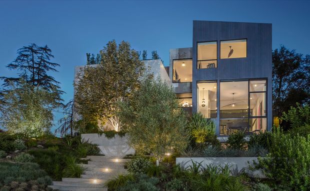 Twilight exterior view of a contemporary multi-storey residence set within a landscaped garden. The home features a tall dark vertical-clad volume, extensive floor-to-ceiling glazing, projecting balconies, and warm interior lighting. Layered concrete retaining walls, steps, ornamental grasses, shrubs, and mature trees form the foreground landscape.