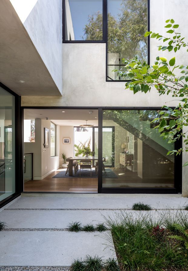 A contemporary two-storey courtyard-facing house with smooth light rendered walls, extensive black-framed glazing, and strong cantilevered geometric forms. The view looks from a landscaped concrete courtyard through large sliding glass doors into a bright dining and living area with timber flooring and restrained furnishings. Trees and grasses soften the minimalist architectural composition.