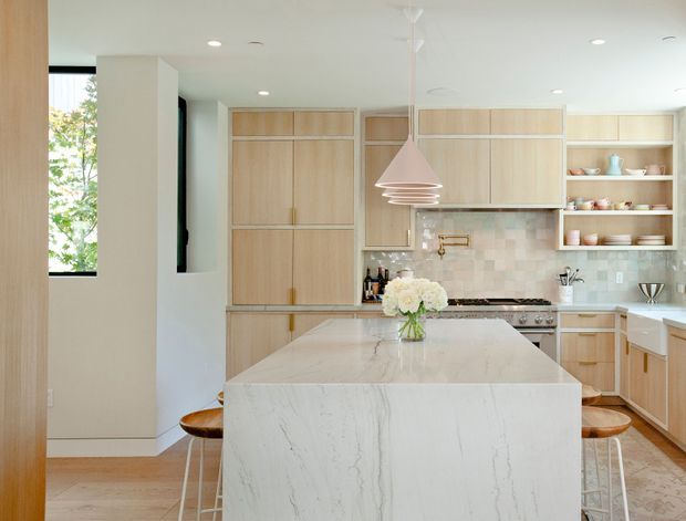 Bright contemporary kitchen with a large waterfall-edge stone island, pale timber cabinetry, open display shelving, a light tiled backsplash, integrated appliances, timber flooring, and sculptural pendant lighting. Bar stools and a floral arrangement sit at the island, while a window provides natural light on the left.