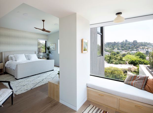 Bright contemporary bedroom with a crisp white upholstered bed, textured vertical wall treatment, pale blue-grey walls, timber flooring, and a built-in window seat with light timber drawers. Large windows overlook a leafy suburban landscape, while a ceiling fan and pendant light provide simple modern detailing.