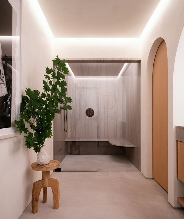 A warm, minimalist bathroom featuring a spacious walk-in shower with frameless glass, pale stone-look wall and floor surfaces, a built-in bench, concealed linear drainage, and a wall-mounted shower fixture. Soft integrated ceiling lighting creates a calm glow. An arched timber door and timber vanity edge add warmth, while a leafy plant on a small sculptural wooden stool provides natural contrast.