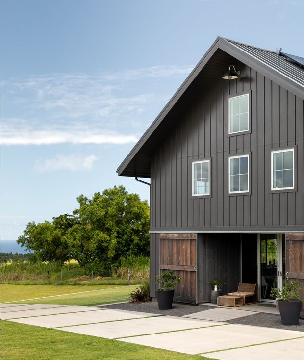 Exterior view of a modern two-storey gable-roof home with dark vertical cladding, a light metal roof, white-framed windows, and a partially open ground-level garage or entry bay. Concrete slab paving leads through a landscaped lawn, with potted plants and a small outdoor seating area at the entrance. Trees and an open rural or coastal-looking landscape are visible beyond the property.