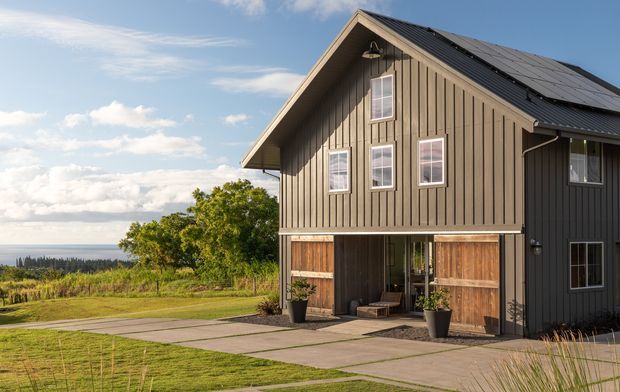 Exterior view of a dark vertical-clad, two-storey gable-roof house on a landscaped rural or elevated site. The building features timber-look garage or barn-style doors, a recessed ground-level entry, white-framed windows, and a large roof-mounted solar panel array. Paved paths and lawn lead toward open countryside and a distant horizon.