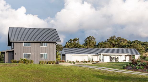 Exterior view of a contemporary rural-style property comprising a tall two-storey gabled building and a long single-storey pavilion. Both buildings have dark grey metal pitched roofs, vertical cladding, and large glazed openings, set within a broad landscaped lawn with native planting and a paved path. Cloudy sky and mature trees form the background.