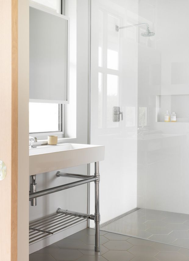 Bright contemporary bathroom viewed through an open timber-framed doorway. The room features a wall-mounted rectangular mirror, a white floating basin with exposed polished metal plumbing and an open lower shelf, a large window, glossy white wall surfaces, a walk-in shower with chrome rain shower hardware and recessed shelving, and pale grey hexagonal floor tiles.