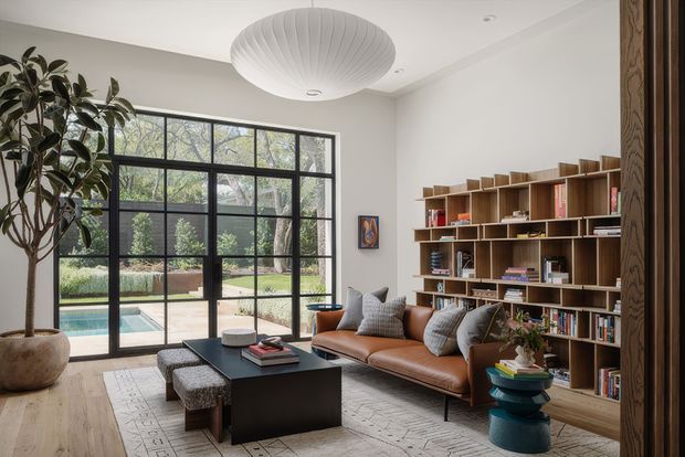 A refined contemporary living room with a large black-framed glazed wall opening to a garden and swimming pool. The room features a low brown leather sofa, black rectangular coffee table, upholstered stools, a large patterned rug, an extensive built-in timber bookshelf, sculptural pendant lighting, indoor planting, and light timber flooring. The space is bright, warm, and carefully curated, with a strong indoor-outdoor connection.