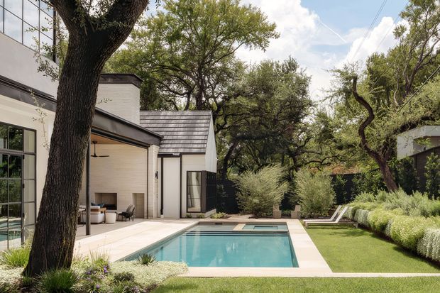 A landscaped residential courtyard viewed from outdoors, featuring a modern white-rendered house with dark metal-framed glazing and a pitched roof, a rectangular swimming pool with pale paving, manicured lawn, mature trees, ornamental planting, and a covered outdoor entertaining area.