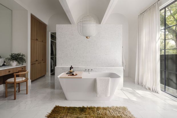 Bright, refined bathroom with a freestanding white bathtub centered in front of a textured white tiled partition. The room features pale marble-look flooring, warm timber cabinetry, a double-height or high vaulted ceiling with curved detailing, a sculptural pendant light, sheer full-height curtains, and large black-framed glazing. A small timber vanity or console with a round mirror is visible to the left, with a textured rug in the foreground.