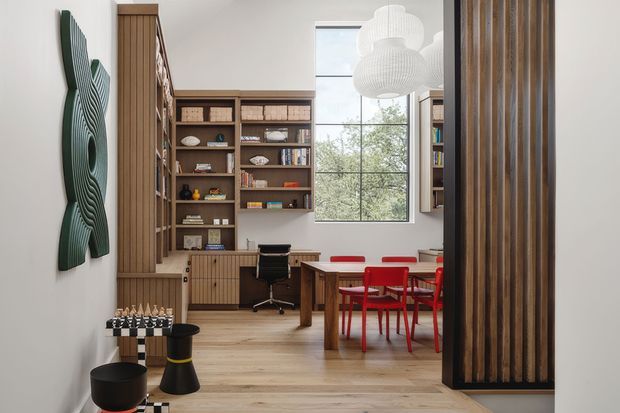 A bright, contemporary open-plan interior combining a home office and dining area. The room features warm timber flooring, extensive built-in timber shelving and cabinetry, a compact desk with an office chair, and a timber dining table surrounded by vivid red chairs. A tall window provides natural light, while a vertical timber slatted divider frames the right side. Sculptural pendant lights and wall art add decorative detail.