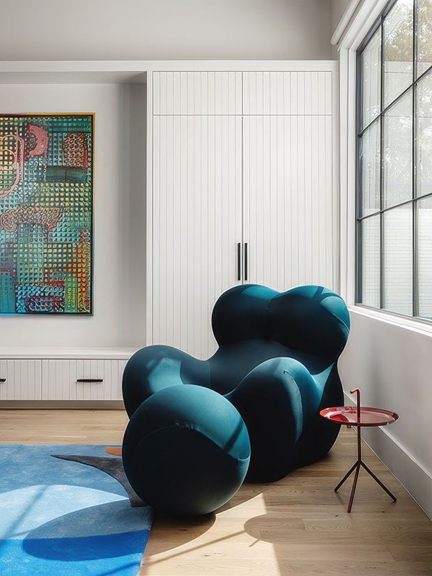 A bright, contemporary sitting area with a sculptural deep teal lounge chair, a small red side table, pale timber flooring, white built-in cabinetry with vertical panel detailing, and a large black-grid window. A colourful abstract artwork and blue patterned rug add bold accents, while strong natural light creates a crisp, inviting atmosphere.