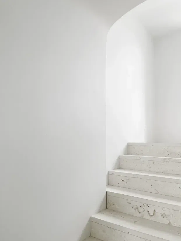 Minimalist interior stairway with broad pale stone steps ascending along a smooth white wall to a softly rounded archway. The scene is bright, monochromatic, and sparsely detailed, emphasizing clean lines and sculptural curves.