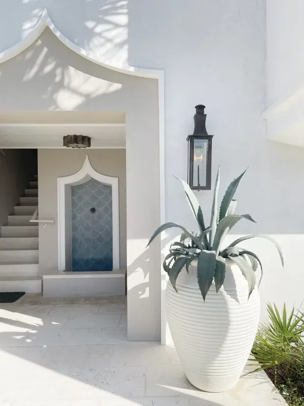 Bright white Mediterranean-inspired courtyard or entryway with sculptural arched detailing, a small decorative blue scallop-tiled fountain niche, pale stone flooring, and a large ribbed white planter containing an agave. A dark lantern is mounted above the planter, while an adjacent stairway leads upward through the arched opening. Strong sunlight creates patterned shadows across the white walls and floor.