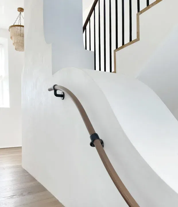 Bright minimalist interior view focused on a sculptural white staircase with smooth curved plastered sides, a timber handrail with dark metal brackets, and contrasting black vertical balustrade rails above. Pale timber flooring and a woven pendant light are visible in the adjacent space.