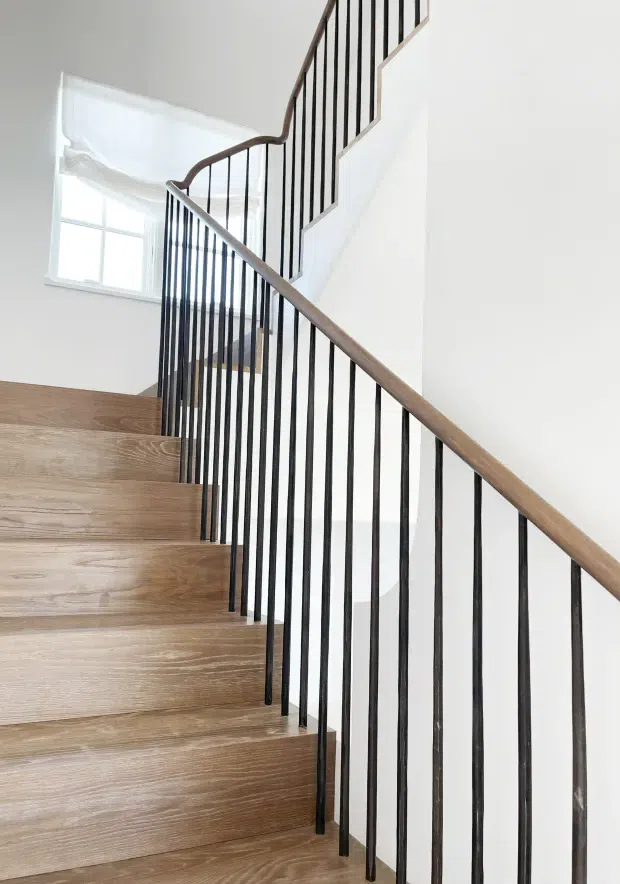 Bright interior view looking up a modern staircase with broad light timber treads, white walls, and a slim black vertical metal balustrade topped by a warm timber handrail. A large window brings in natural light at the upper landing.