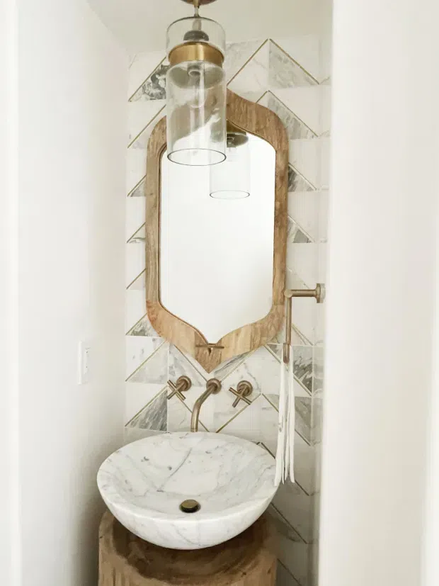 A compact bathroom powder room featuring a round white marble vessel sink on a rustic timber pedestal, wall-mounted aged brass tapware, and a large irregularly framed timber mirror. The wall is clad in geometric patterned marble-look tiles, with a hanging glass-and-brass pendant light reflected in the mirror. The palette is warm, neutral, and natural with a refined eclectic character.