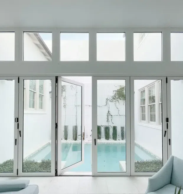 Bright, minimalist interior looking through a wall of white-framed glazed doors and upper clerestory windows onto a small turquoise courtyard pool. The room contains pale upholstered seating at the lower edges, while the enclosed courtyard has white walls, climbing greenery, and a clean contemporary feel.
