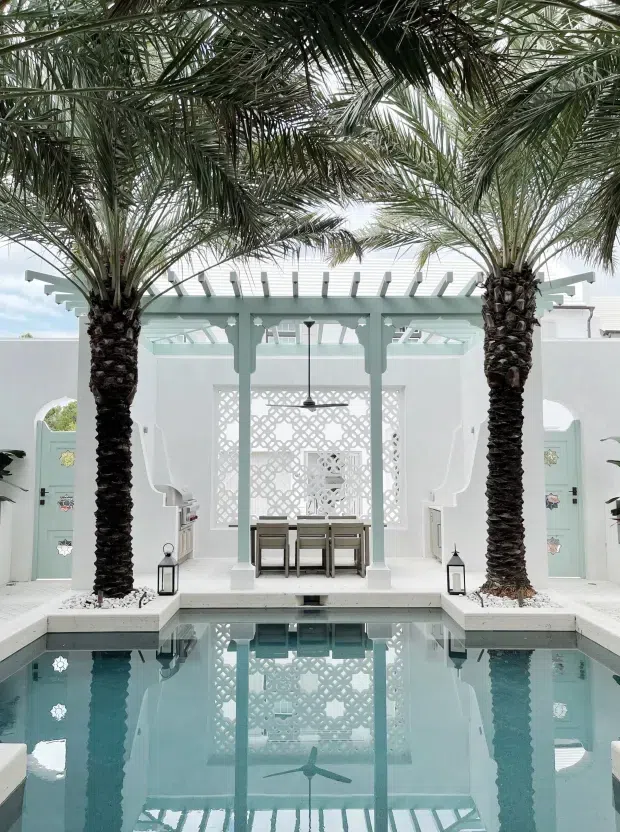 A bright, symmetrical courtyard pool framed by two mature palm trees. A pale aqua and white pavilion sits beyond the pool, with arched openings, decorative breeze-block screening, pergola-like roof beams, glass roof panels, and a dining table beneath a ceiling fan. The pool reflects the pavilion and palms, creating a serene resort-like outdoor setting.