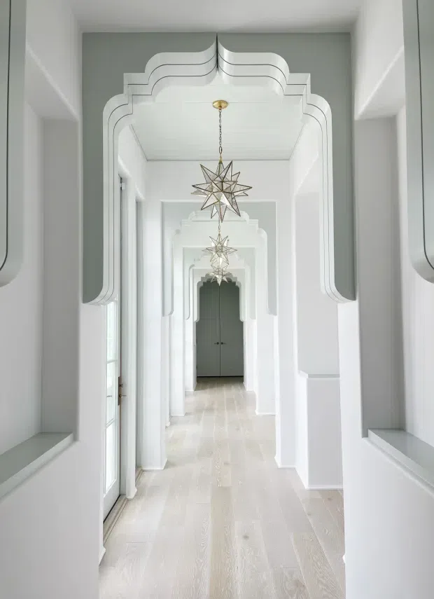 A bright, elegant hallway with a series of repeated white ornamental archways and layered geometric wall mouldings leading toward dark double doors. Pale timber flooring, white panelled walls, glazed side doors, and a sequence of suspended star-shaped pendant lights create a refined, symmetrical perspective.