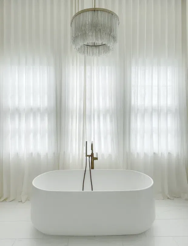 A bright, serene bathroom centered on a white freestanding oval bathtub. Sheer white floor-to-ceiling curtains cover the windows, creating a soft diffused backdrop, while a brass-toned tub filler and a layered fringed pendant light provide subtle contrast. The floor is finished in white tile.