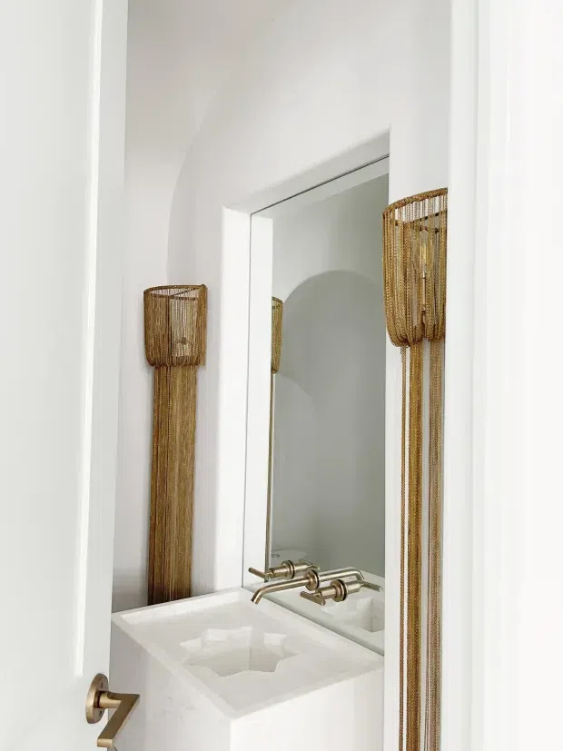 A narrow, bright white bathroom or powder room viewed through a partially open white door. The space features a rectangular integrated white basin with wall-mounted brushed metal tapware, a large frameless mirror, and distinctive tall woven pendant or wall lights in warm natural brown tones. The styling is clean, refined, and minimalist with subtle sculptural forms.