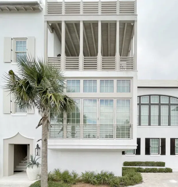 A bright white multi-storey residential facade with large glazed windows, dark shutters, a projecting enclosed balcony, and a distinctive upper-level open porch with louvred privacy screens and exposed timber framing. Tropical palms, clipped hedges, and a paved entry garden frame the building.