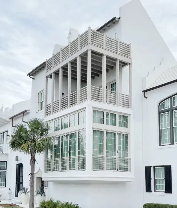 Exterior view of a bright white multi-storey coastal-style building with prominent enclosed glass rooms, tall gridded windows, dark shutters, horizontal balcony railings, slender white columns, and a projecting upper-level terrace. A palm tree and potted landscaping sit in front of the facade.