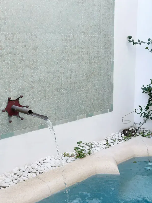 Close-up architectural detail of a sculptural bronze-toned wall spout pouring water into a small blue-tiled pool or water feature. The backdrop is a pale textured mosaic tile feature wall bordered by narrow green-grey tiles, with white decorative river stones and sparse planting along the pool edge. The scene has a clean, serene courtyard or spa-like character.