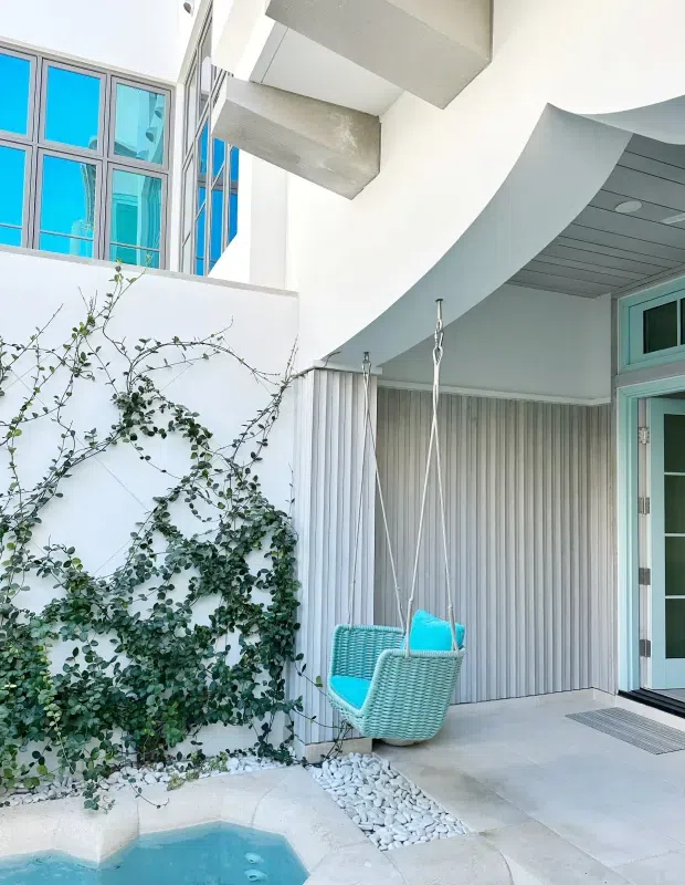 Bright contemporary courtyard or covered outdoor entry area with white curved architectural forms, turquoise-framed glazing, a ribbed vertical wall finish, a hanging woven chair with vivid cyan cushions, climbing greenery, white pebble landscaping, and the edge of a small blue pool or spa.