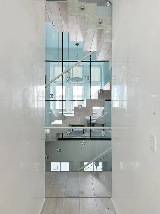 A narrow interior view through a tall full-height mirror, reflecting a bright multi-level stairwell and open-plan dining area. The mirror captures floating or sculptural pale timber stair elements, a slim handrail, expansive black-framed glazing, pale timber flooring, a dining table with upholstered chairs, and multiple spherical pendant lights. The scene is light, airy, and contemporary with strong vertical layering and reflective surfaces.