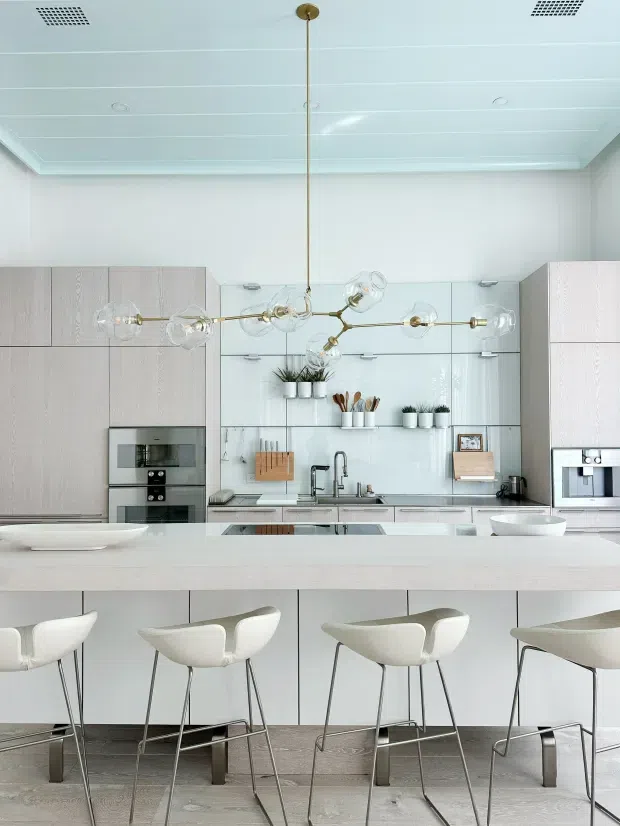 Bright contemporary kitchen with pale timber cabinetry, a large light island with integrated seating, white engineered-stone surfaces, mirrored splashback shelving, integrated appliances, and a sculptural brass multi-globe pendant light. The space has a clean, refined, softly neutral appearance.