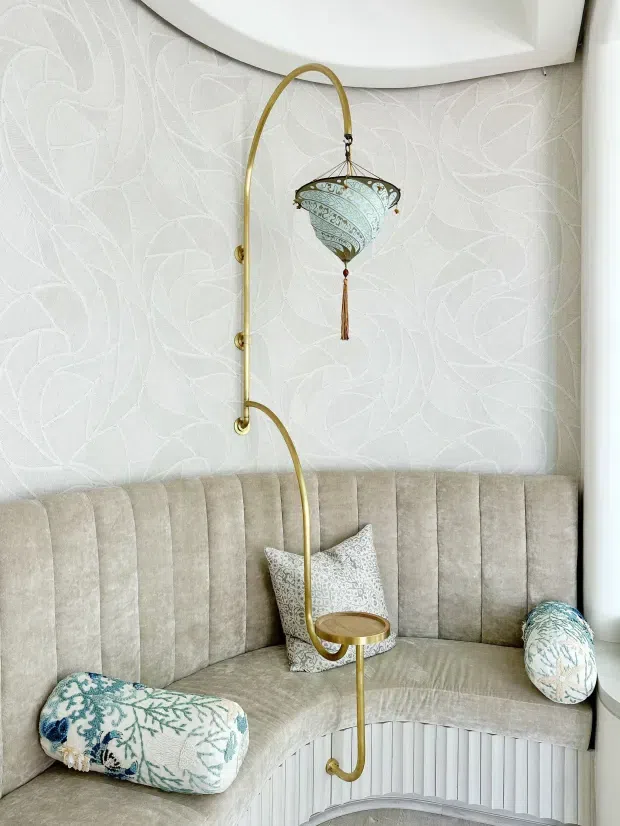 Close-up of a curved built-in upholstered banquette beneath a large rounded white architectural feature, with vertical channel tufting, pale botanical patterned wallcovering, decorative cushions, and an ornate brass arched floor lamp with a hanging patterned shade and integrated small table. The palette is soft ivory, beige, pale blue, and brass, creating an elegant decorative sitting nook.