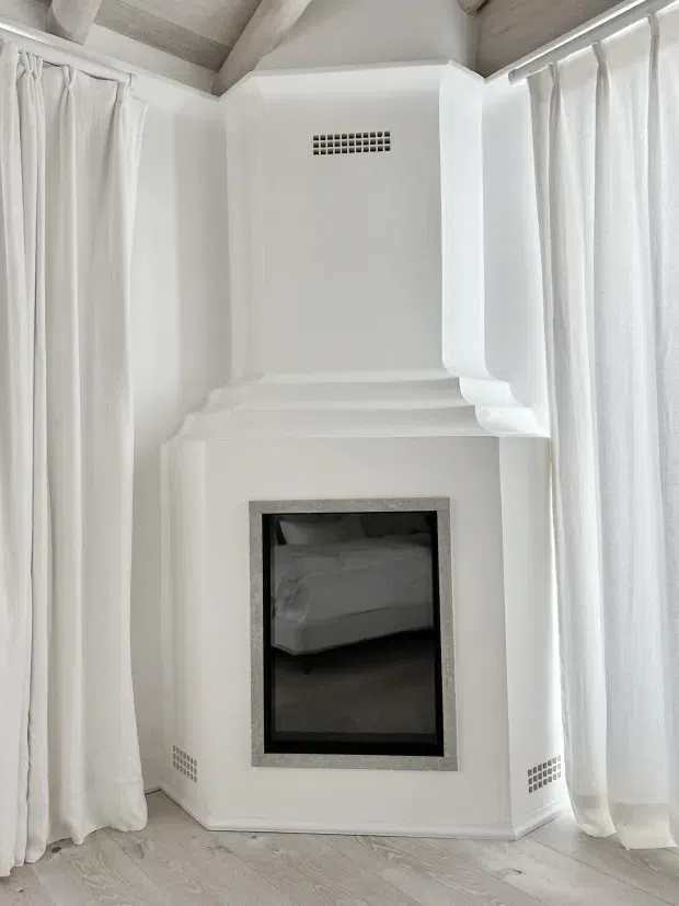 A bright white bedroom corner with a tall, ornate fireplace surround and dark firebox. Floor-to-ceiling sheer white curtains frame the corner on both sides, while pale timber-look ceiling beams and light timber flooring add subtle texture. The fireplace opening reflects a bed, confirming the room as a bedroom.