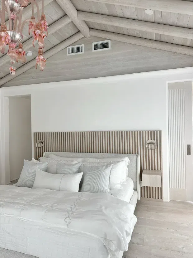 Bright, softly styled bedroom with a white upholstered bed, layered white bedding and pillows, integrated ribbed wood headboard with matching bedside niche, and wall-mounted reading lights. The room features pale timber flooring, white walls, a vaulted ceiling with exposed pale timber beams and paneling, vertical white wall detailing, and a delicate pink glass chandelier.