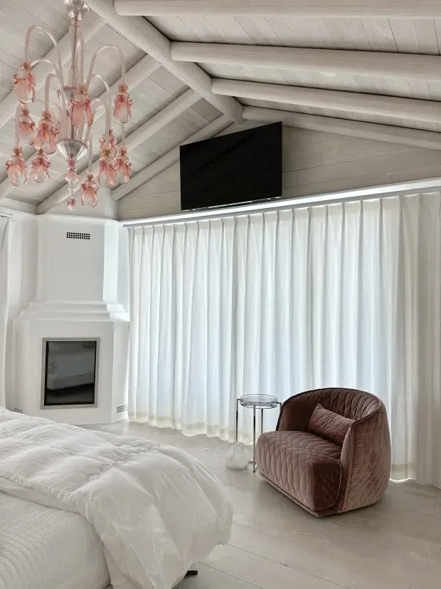 A bright, predominantly white bedroom with a pitched ceiling featuring exposed painted timber beams and planks. Floor-to-ceiling sheer white curtains span the wall, beside a sculptural white fireplace with a dark firebox. The room includes a white bed, a plush blush-brown quilted armchair, a small round side table, a wall-mounted television, and a delicate pink glass chandelier.