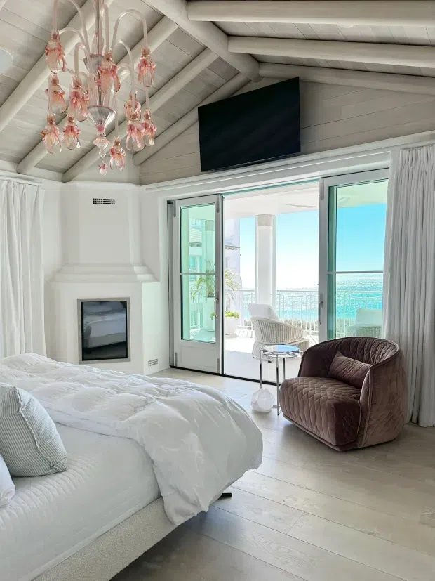 Bright coastal bedroom with a white upholstered bed, exposed white-painted timber rafters, pale timber flooring, a sculptural pink glass chandelier, fireplace, wall-mounted television, and floor-to-ceiling glazed doors opening to a balcony with ocean views. A plush brown lounge chair sits beside the glazing.