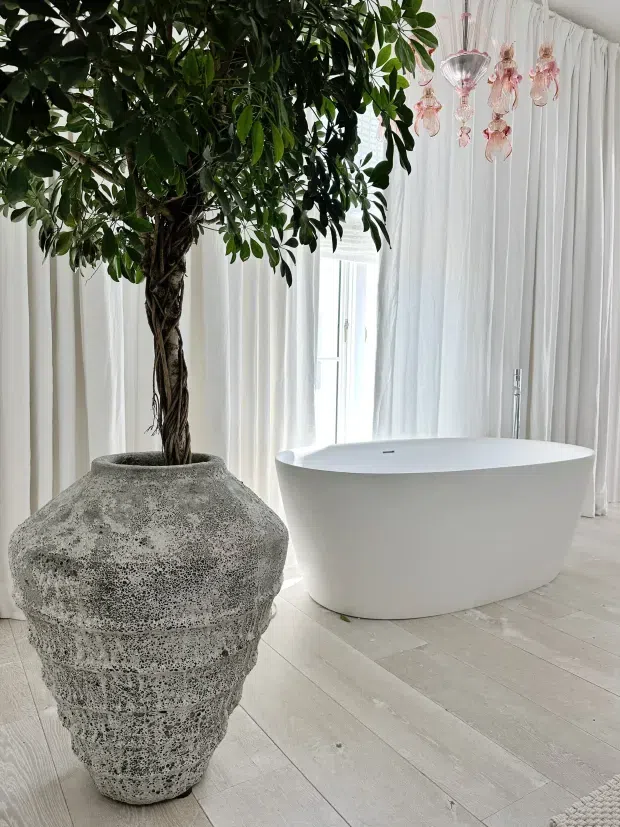 A bright, serene bathroom featuring a freestanding white bathtub beside full-height sheer white curtains. A large textured grey stone planter with a mature indoor tree forms a prominent natural feature, while delicate pink hanging glass lights add a decorative accent. Pale timber-look flooring and predominantly white finishes create an airy, spa-like atmosphere.