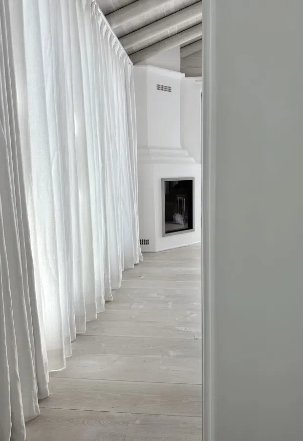 A bright, minimalist interior viewed through a doorway or narrow opening. Sheer white curtains extend along the left side, while pale whitewashed timber flooring and a white built-in fireplace with a black firebox dominate the space. The ceiling features exposed pale timber boards or beams, creating a softly textured architectural detail. The overall composition is calm, monochromatic, and highly pared back.