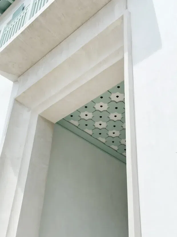 Close-up architectural view of a pale, heavily textured concrete or plaster-framed opening beneath a projecting upper level. The recessed soffit features a repeating mint-green and white geometric patterned surface with small dark circular accents. A white balustrade is partially visible at the upper left, and the overall composition is light, minimal, and detail-focused.