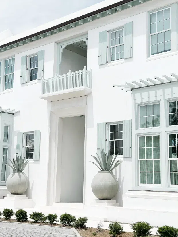 Bright white multi-storey residential facade with pale blue-green shutters, divided-light windows, decorative balcony railing, a tall central entry opening, and projecting pergola-like sunshades. Symmetrical classical detailing and large round planters with architectural succulents create a refined coastal character.