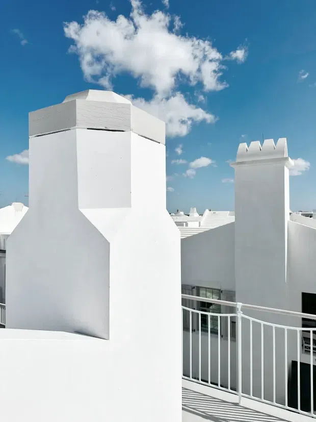 Exterior architectural photograph of a cluster of bright white, flat-roofed buildings with tall parapets and distinctive crenellated chimney caps. A white metal balcony or roof terrace railing occupies the foreground, with additional white roof forms visible beyond. The scene is strongly sunlit beneath a vivid blue sky with scattered clouds.