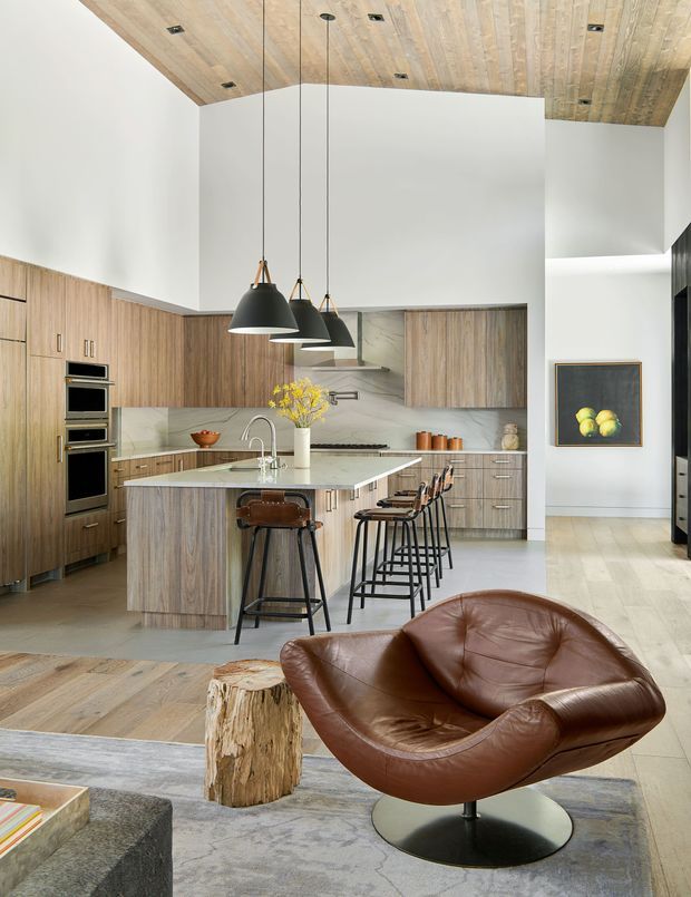 Open-plan contemporary kitchen and living area with extensive vertical wood cabinetry, a large waterfall-edge island, integrated appliances, pale stone surfaces, and a dramatic wood-plank ceiling. A row of black pendant lights hangs above the island, while a brown leather lounge chair and rustic timber stump table occupy the foreground.