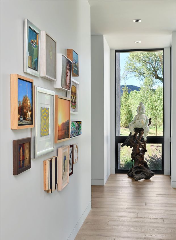 A bright modern interior hallway or gallery passage with a curated collection of framed artwork arranged across a pale wall. The corridor has recessed ceiling downlights, light timber plank flooring, crisp white walls, and a tall black-framed glazed door or window at the end overlooking landscaped greenery. A sculptural organic wood-and-stone artwork stands in front of the glazing.