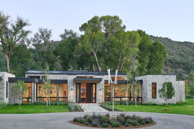 Single-storey contemporary residence set against a wooded, sloping landscape. The low horizontal composition combines pale stone masonry, dark metal roofing, timber-toned cladding and extensive floor-to-ceiling glazing. A covered entry with projecting flat canopy opens onto a patterned paved path, with manicured lawn and young trees in front.