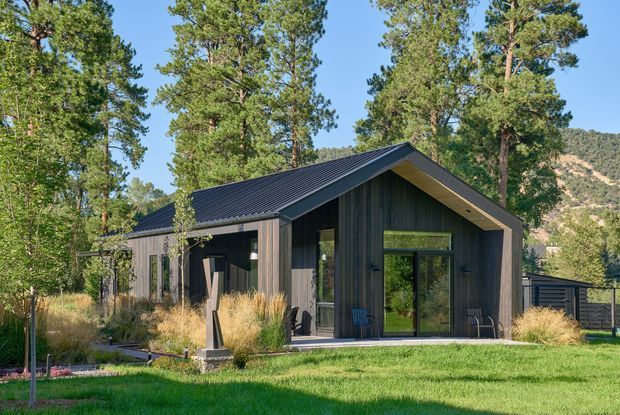 A modern single-storey cabin-style house set within a landscaped lawn and tall pine trees. The building has a simple gable roof, dark vertical timber cladding, deep charcoal metal roofing, and large black-framed glazed doors and windows. Natural grasses and a mountain backdrop reinforce the wooded setting.