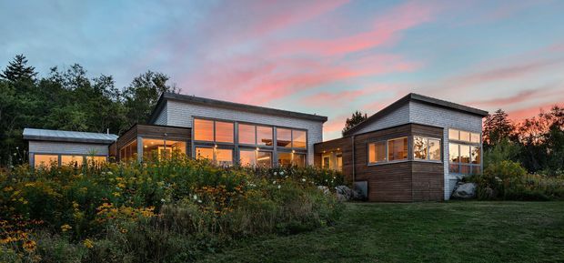 Single-storey contemporary residential house viewed from the garden at dusk, composed of several connected dark timber-clad volumes with low-pitched roofs and extensive warm-lit glazing. Native-style planting and meadow garden surround the building, with a broad lawn in the foreground and a colourful sunset sky behind.