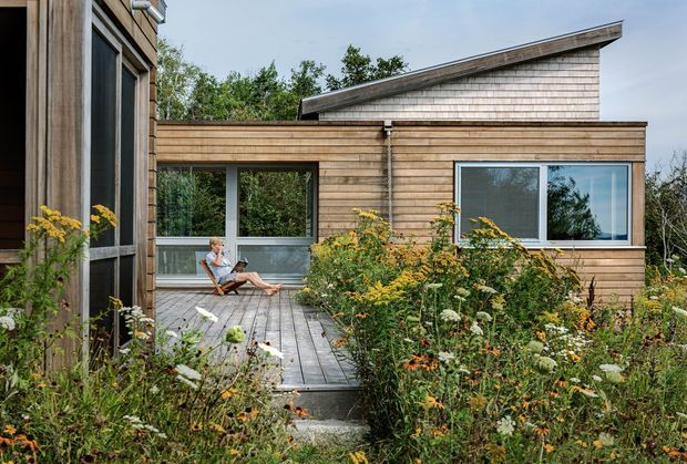 Exterior view of a contemporary timber-clad house arranged around a planted courtyard and timber deck. The low horizontal wing has large windows and a glazed corner, while a taller section behind features a distinctive sloping roofline. Native-style garden planting surrounds the deck, with one person seated outdoors.