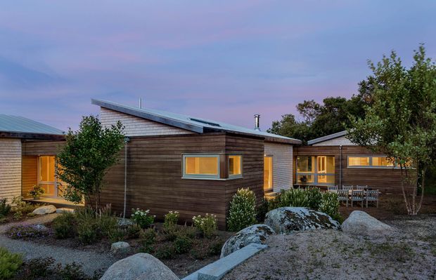 Exterior view of a low, single-storey cluster of timber-clad buildings arranged around a landscaped gravel garden. The structures have simple shed and low-pitched roof forms, warm illuminated windows, and a restrained contemporary cabin-like appearance. Large natural boulders, native-style planting, and gravel define the foreground.