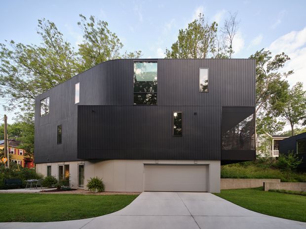 Exterior view of a contemporary multi-level residential building with a boxy cantilevered form. The upper storey is clad in dark vertically ribbed metal, with irregularly placed windows and a projecting glazed corner. The lower level has a pale rendered finish, garage door, entry, lawn, and concrete driveway, with mature trees and neighbouring houses around the site.