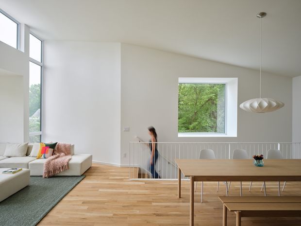 Bright open-plan interior combining a living room and dining area, with white walls, extensive glazing, pale timber flooring, a neutral sectional sofa, and a timber dining table with bench seating. A stairwell with a slim white balustrade connects the spaces, and a sculptural pendant light hangs above the dining table. Greenery is visible through the windows.