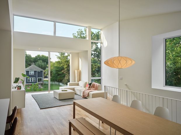 Bright open-plan interior combining a dining area and living room, with a long light-wood dining table and bench in the foreground, a pale sofa and ottoman beyond, and expansive floor-to-ceiling and clerestory glazing overlooking greenery and a neighbouring house. A sculptural pendant light hangs above the dining table, while a white stair balustrade separates the levels. The space has a clean, contemporary design with abundant natural light and high ceilings.
